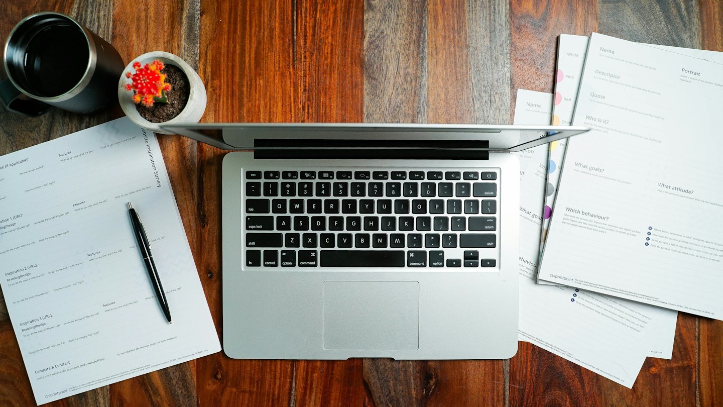 Laptop, Umfragedokumente und Stift auf einem Holztisch.